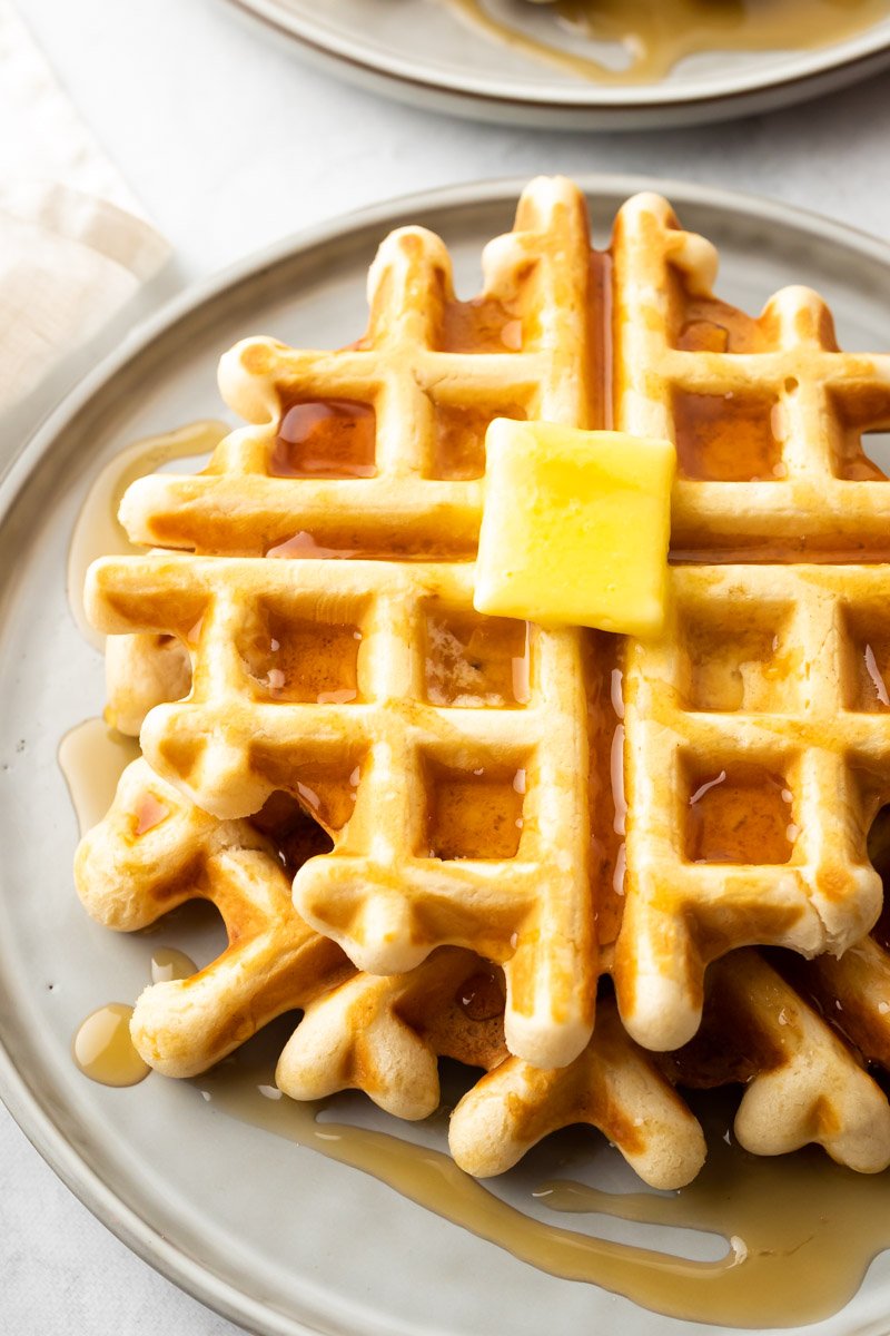 Nahaufnahme eines Stapels Waffeln auf einem beigen Teller, mit Sirup übergossen und mit einem Stück Butter belegt