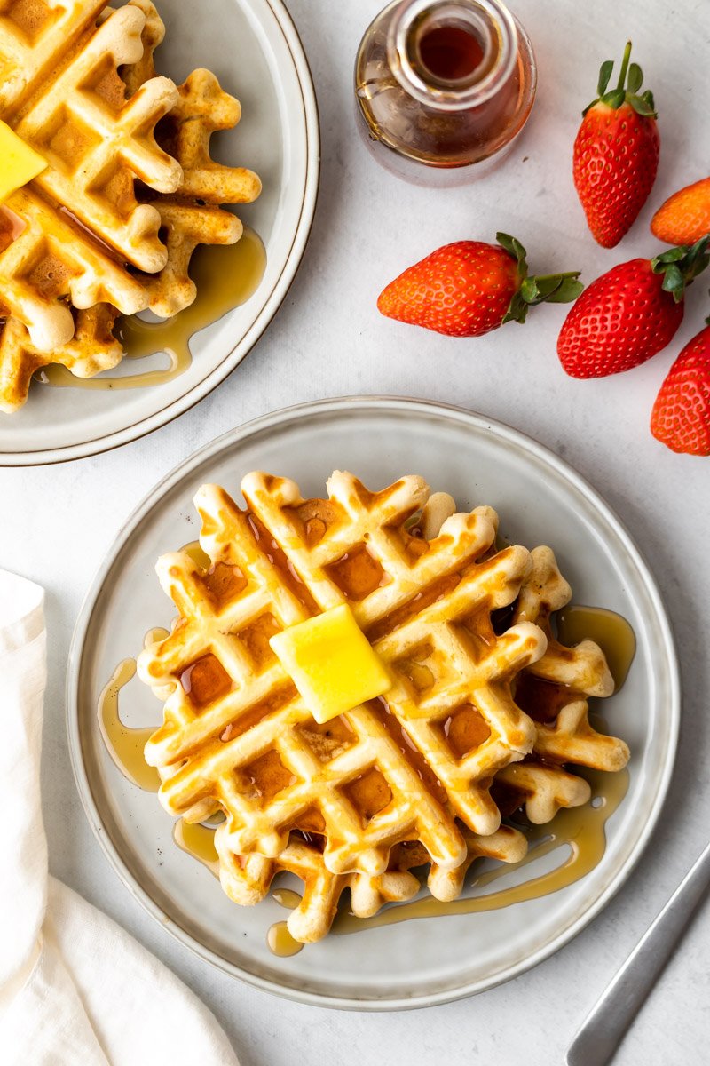 Draufsicht auf zwei Teller mit Waffeln neben ganzen Erdbeeren und einem Glas Sirup