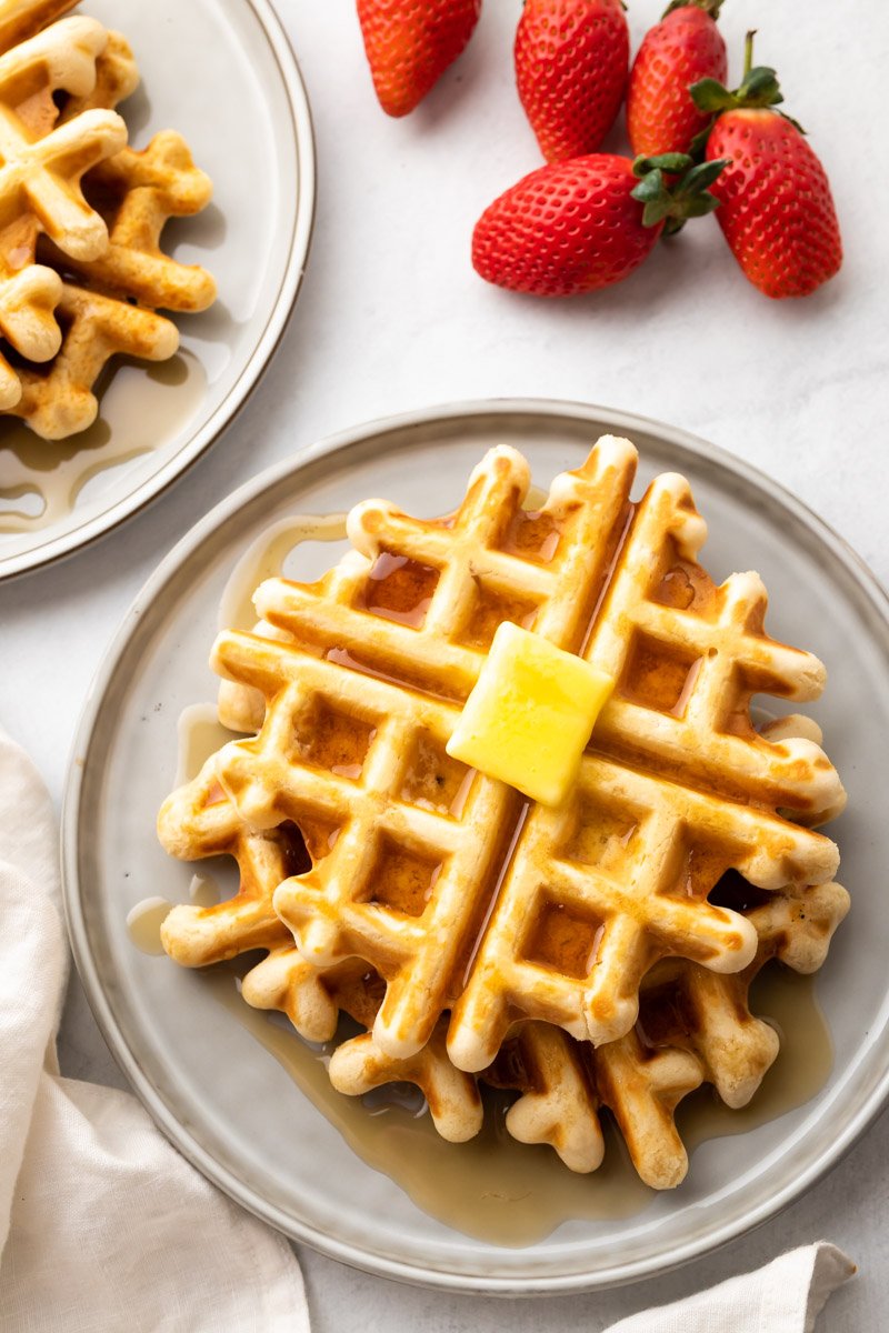 Draufsicht auf zwei Teller mit Waffeln, die mit Sirup übergossen und mit Butter belegt sind, neben ganzen Erdbeeren